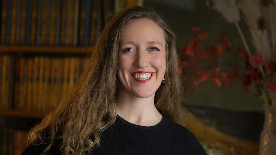 A women with long hair, wearing black smart attire, smiling at the camera, with a wood-panelled background behind her.